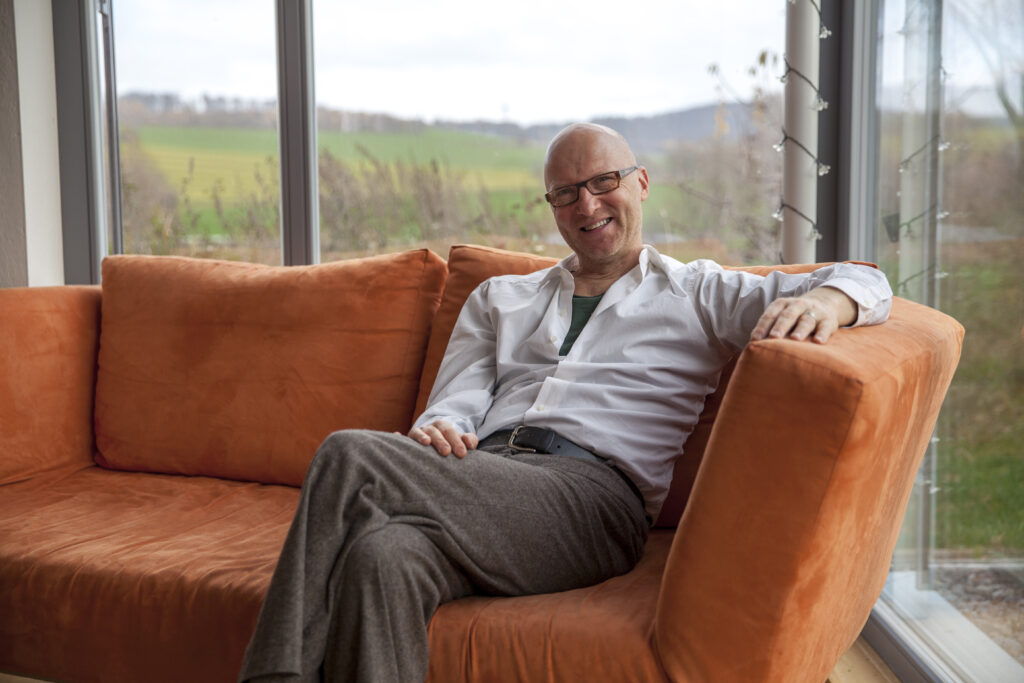 Manfred is sitting on a organge sofa smiling at the camera. Manfred is a toll man, bolg, with glasses. It's really elegant. 